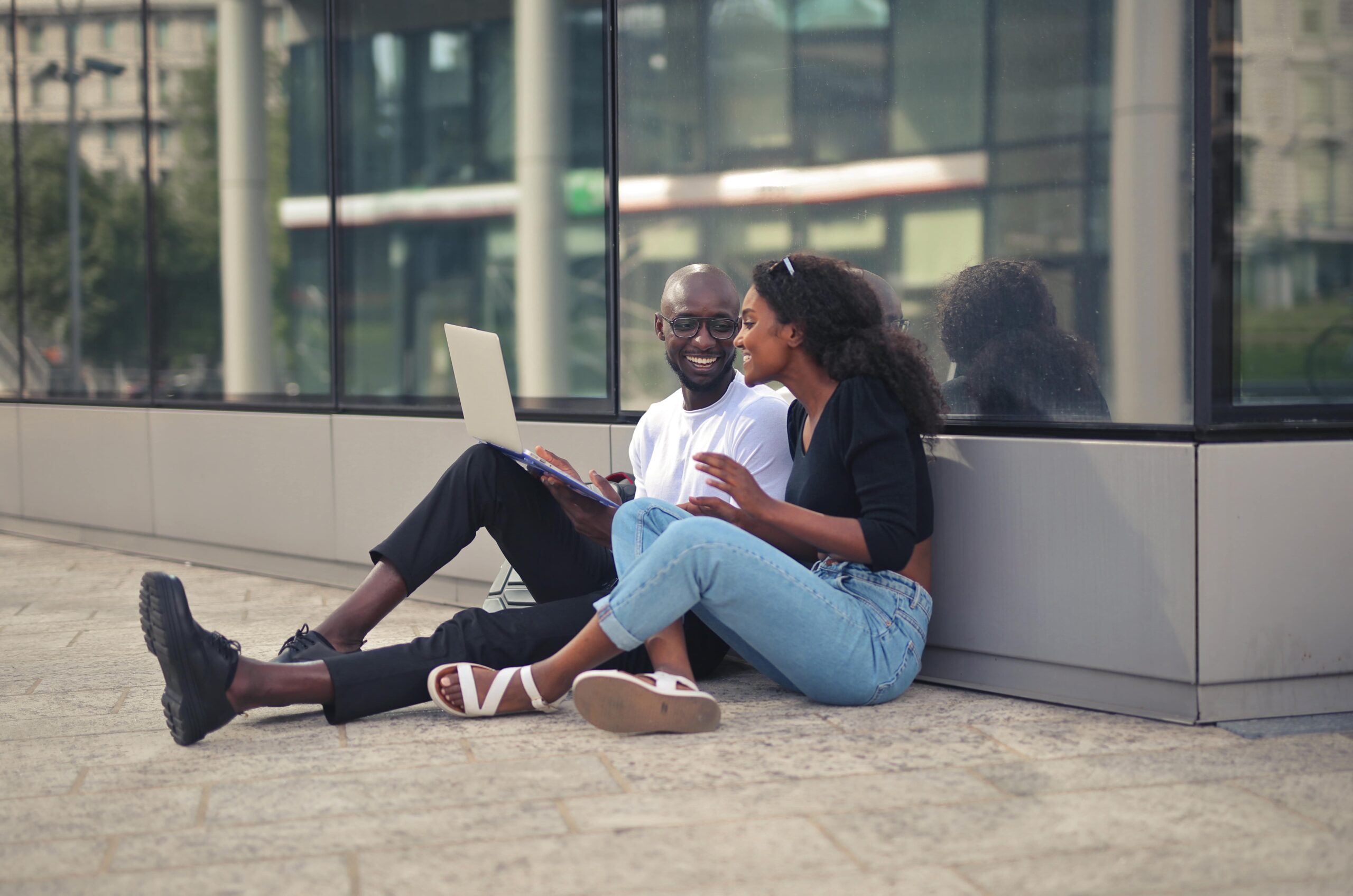 cheerful-smiling-african-male-female-sitting-ground-using-laptop-daytime-min